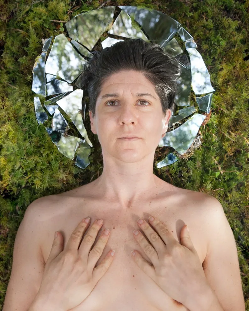 Human bust with hands over chest area, eyes look straight out at viewer with head against a broken mirror circle