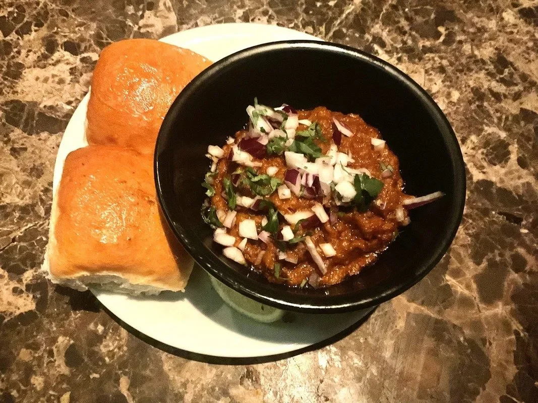 A bowl with a spicy mashed vegetables, some fresh onion pieces sprinkled over it, and two bread buns to the side