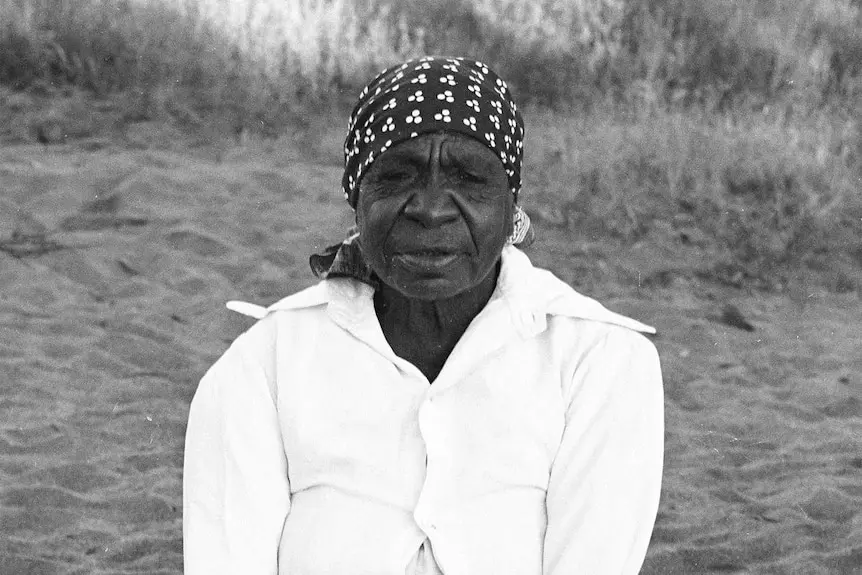 A portrait of artist Emily Kam Kngwarray kneeling on Country wearing a white shirt and head scarf