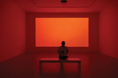 A figure sitting on a bench is silhouetted by a glowing screen that fills the otherwise empty room with orange-red light.
