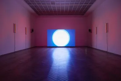 A vast gallery with purple lighting. At its center, on the floor, is a screen showing what looks like a close-up of the sun against a blue sky.