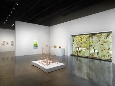 A beige lattice-like sculpture on a low white pedestal centers the gallery. A cluster of prints, a glowing green artwork, two small spherical sculptures, and a waxy window installation are in the background.