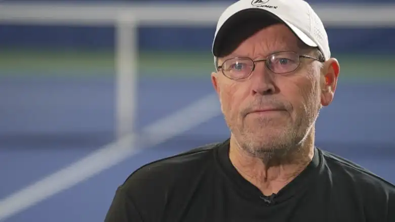 An unsmiling man wearing a ball cap and small circular glasses.