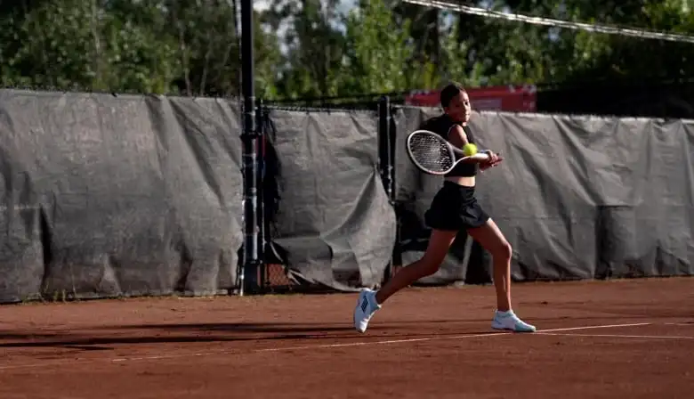 A girl wearing all black on a tennis court with a tennis racket hitting a ball