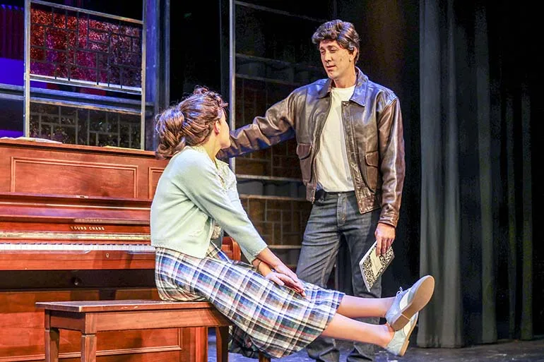 Carole King sits on a piano bench looking up at Gerry in a leather jacket looking like the Fonz