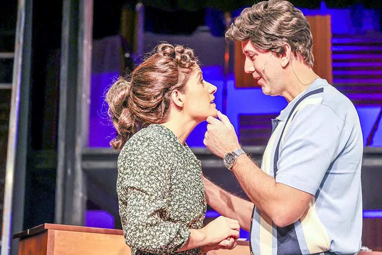 Carole King looks up at Gerry, who is holding her chin and likely singing while wearing a horrible wig