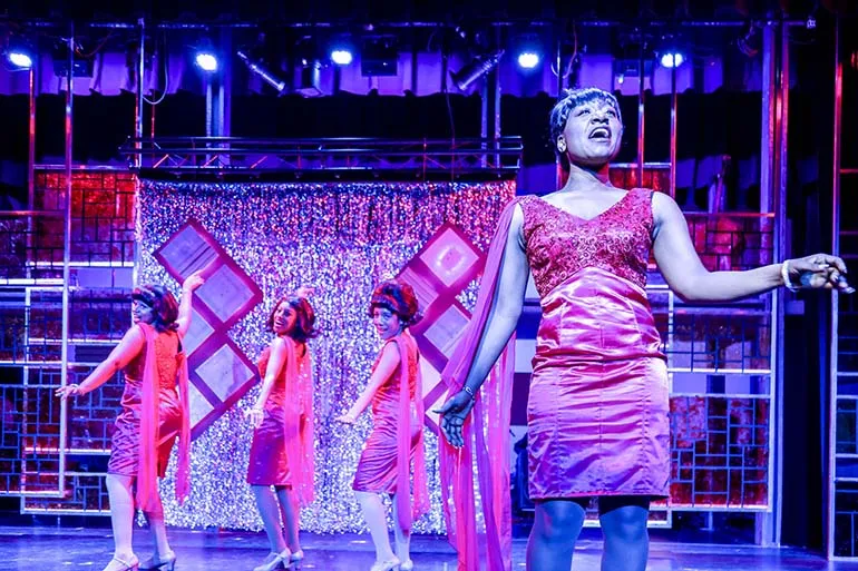 Women in pink dresses belt out songs onstage