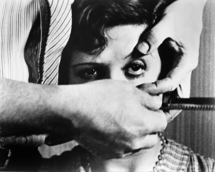 A man with a razor in his hand holds a woman’s eye open