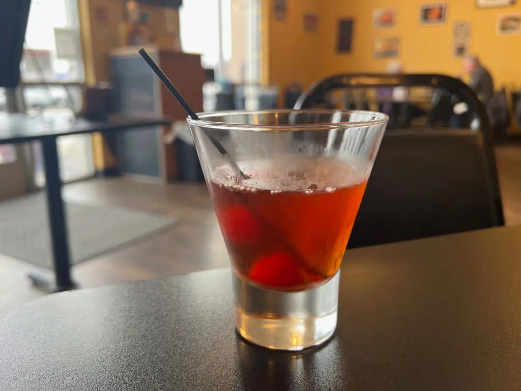 A cocktail called the Manbana which has whiskey and two cherries in a stemless martini glass.