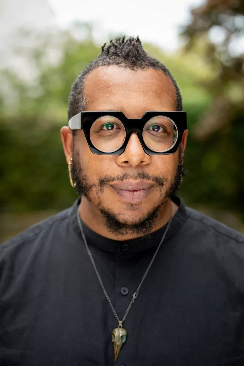 A black man wearing stylish black eyeglasses, gold hoop earrings and a pendant on a chain around his neck on top of his Nehru collar button down shirt smiles at the camera. 