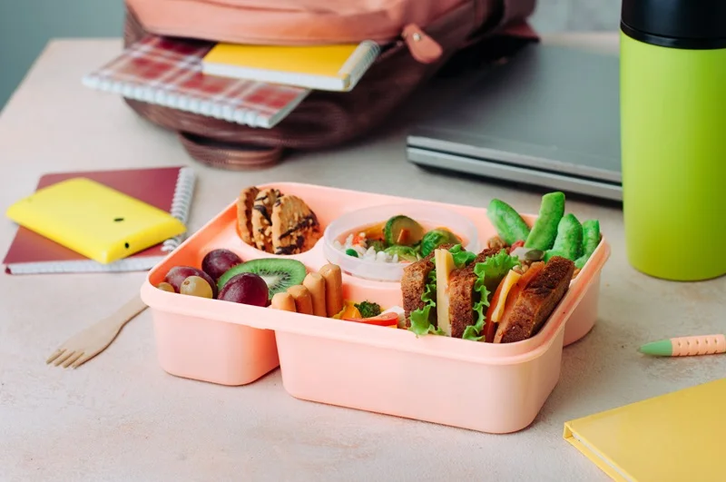 Open lunch box with sandwiches, vegetable salad, fresh grape and kiwi, rice and nuts on the table near backpack, laptop and thermo mug