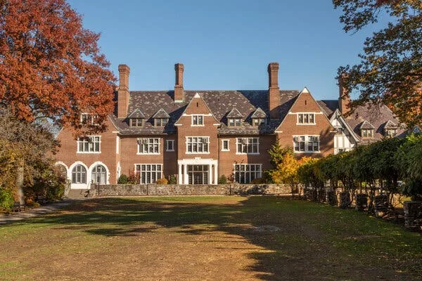 This color photograph shows a stately brick campus building with a long front lawn framed by a low stone wall and trees. 