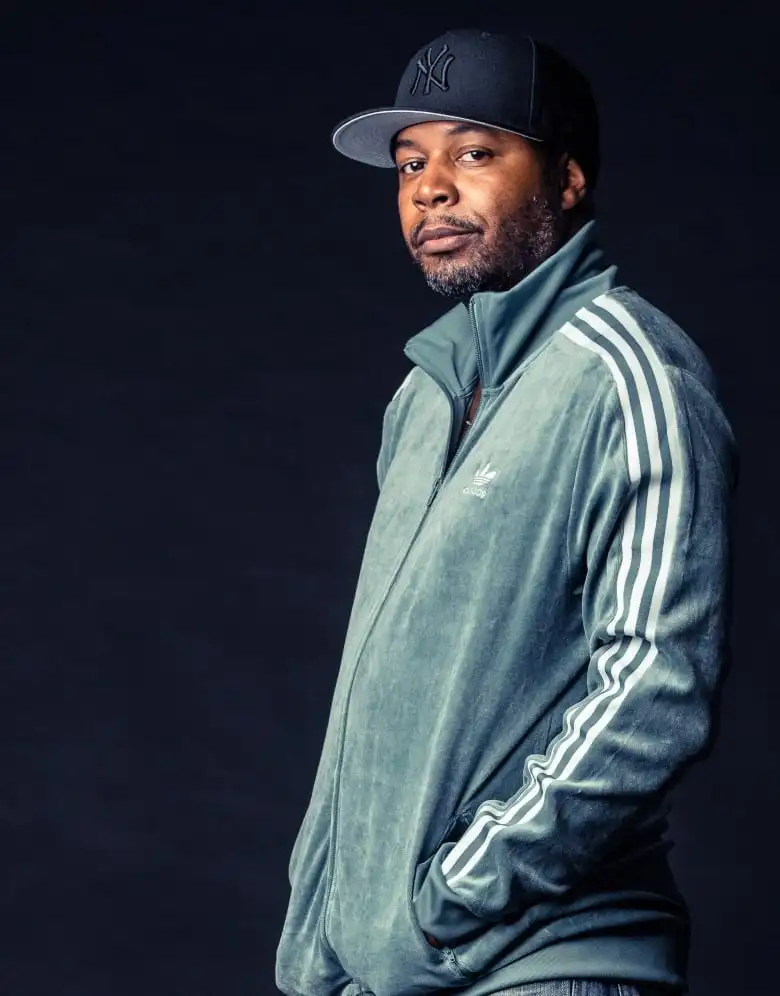 A Black man in a teal Adidas track suit and ballcap stands against a black background. He has his hands in his pockets and is visible from the waist up. He is looking at the camera
