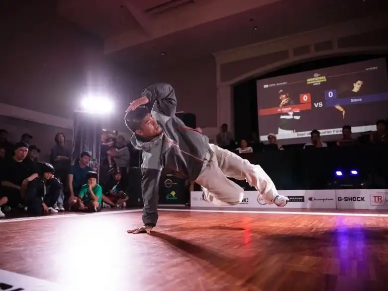 A man break dances in front of a crowd on a wood floor indoors.