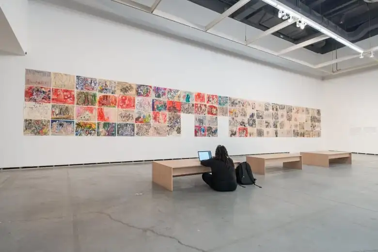 A woman seated on the floor of an art exhibit works on her laptop.
