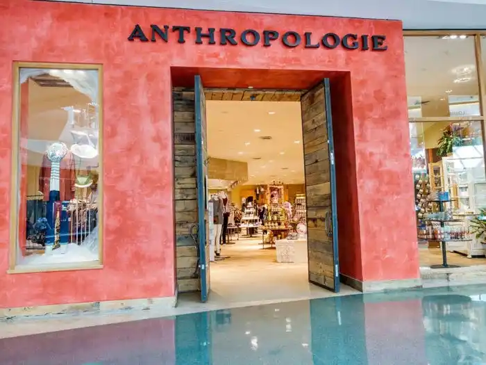 A red-orange Anthropologie storefront with wooden double doors inside of a mall. Clothing and home goods can be seen through the entrance