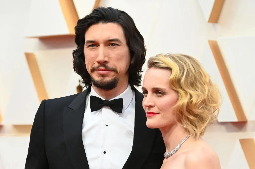 A man in a tuxedo and a woman wearing a jeweled necklace pose on a red carpet