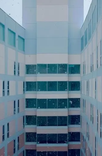 a tall, long rectangular courtyard is shown out of the window of a skyscraper, with other skyscraper walls wrapping around it in shades of reddish brown and gray with black windows. Specs of white scatter across the scene, and could be either specs of dirt on the window or lightly falling snow. A wedge of sky above the building is pale gray. 