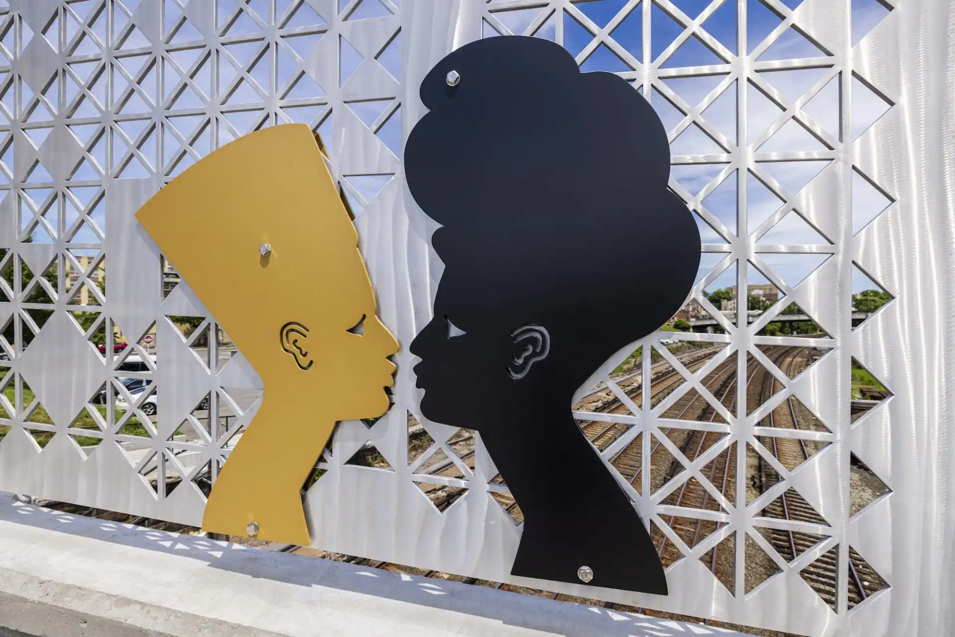 Two metal silhouettes of Nefertiti and a Black woman face-to-face on a bridge above some railroad tracks.