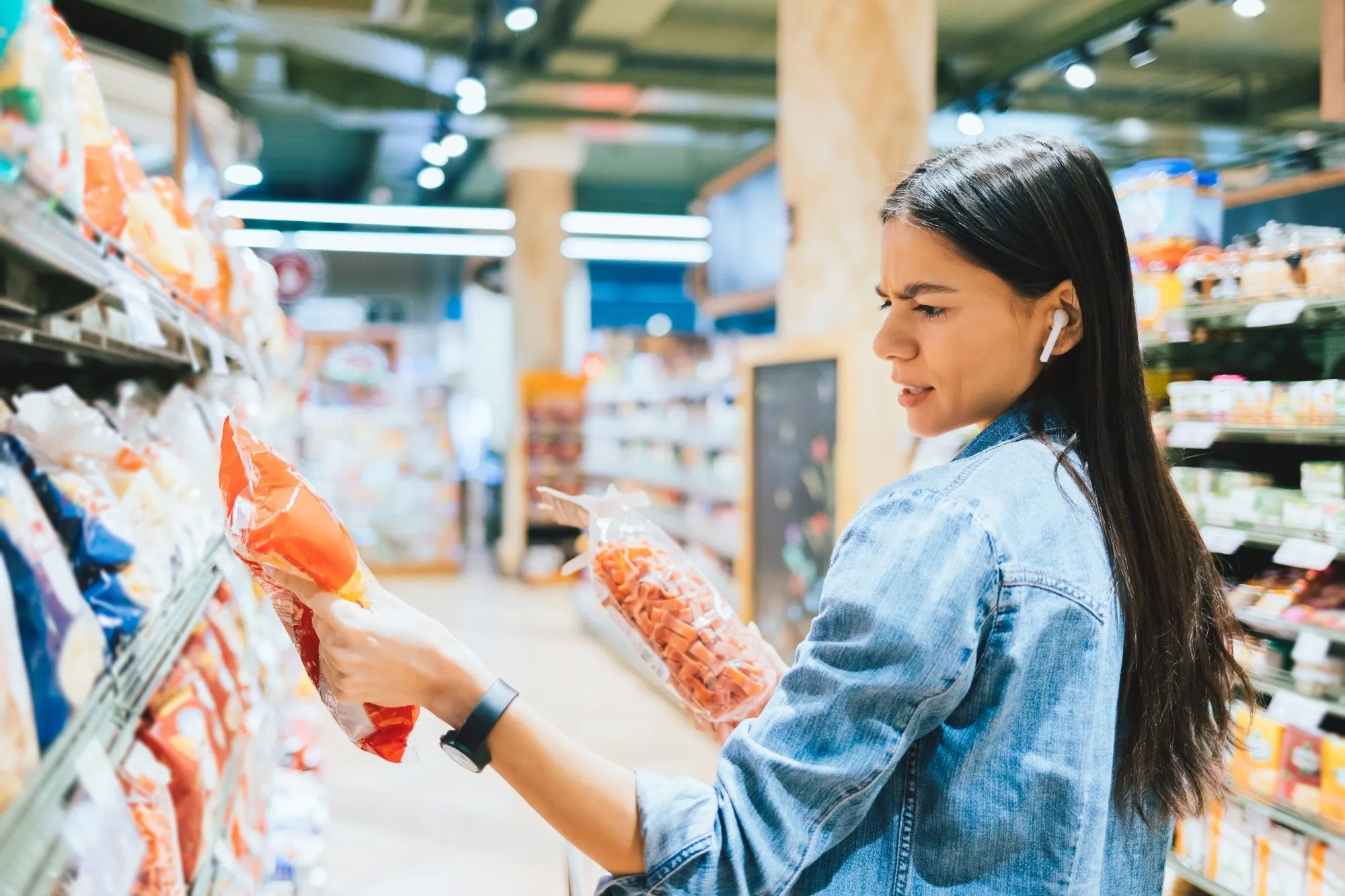 Dissatisfied young woman wearing casual outfit and earphones holding two packs of pasta looking at label while standing in grocery department in supermarket