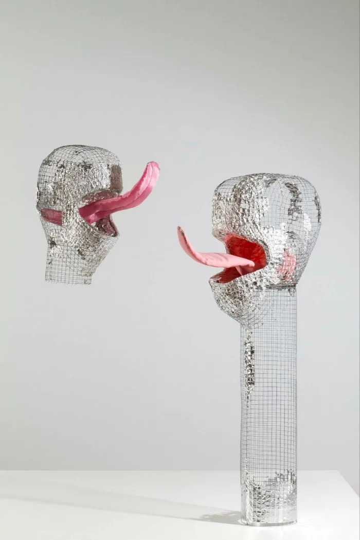Two heads made of steel mesh, partially covered in silver foil, with large pink tongues poking out