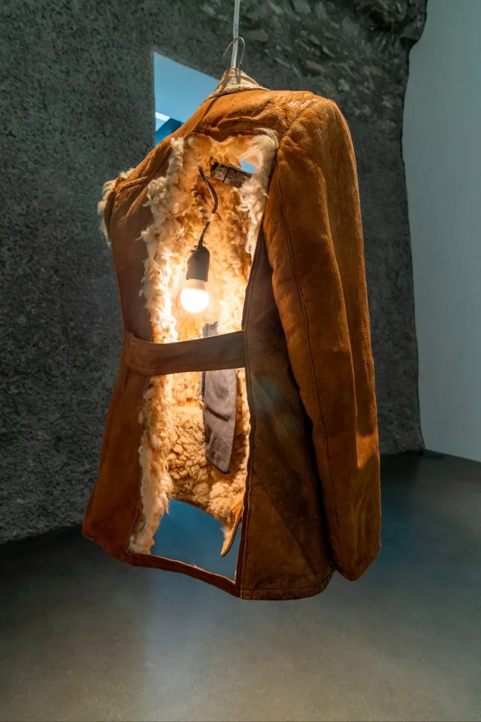 A thick sheepskin coat hanging from a hook with the back cut out and a lit bulb implanted