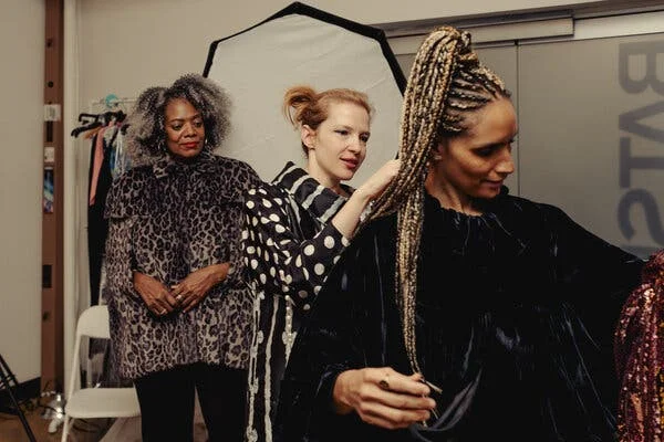 Ms. Hay in a black and white polka-dot dress with her strawberry blond hair pulled back, fits a black velvet dress on a woman with long braids tied atop her head. Behind them, a woman with natural gray hair waits, wearing a faux fur leopard print cape. 