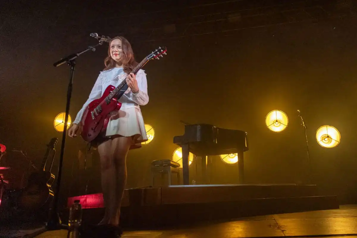 Laufey holding a guitar, with a piano and stage lights in the background