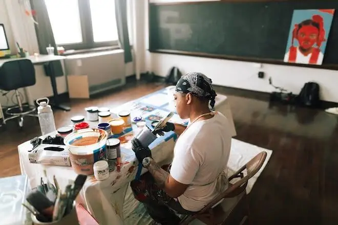 Artist Nazeer Sabree, a Black man wearing a black bandana tied on his head and a white t-shirt, faces away from us as he works at a table covered with paints n his studio. In the distance: a portrait of what appears to be a young girl, all in red. Photo by Guarida.