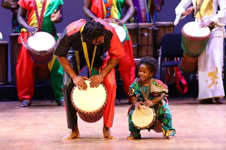 An adult and toddler, both holding drums, perform with the KanKouran West African Dance Company