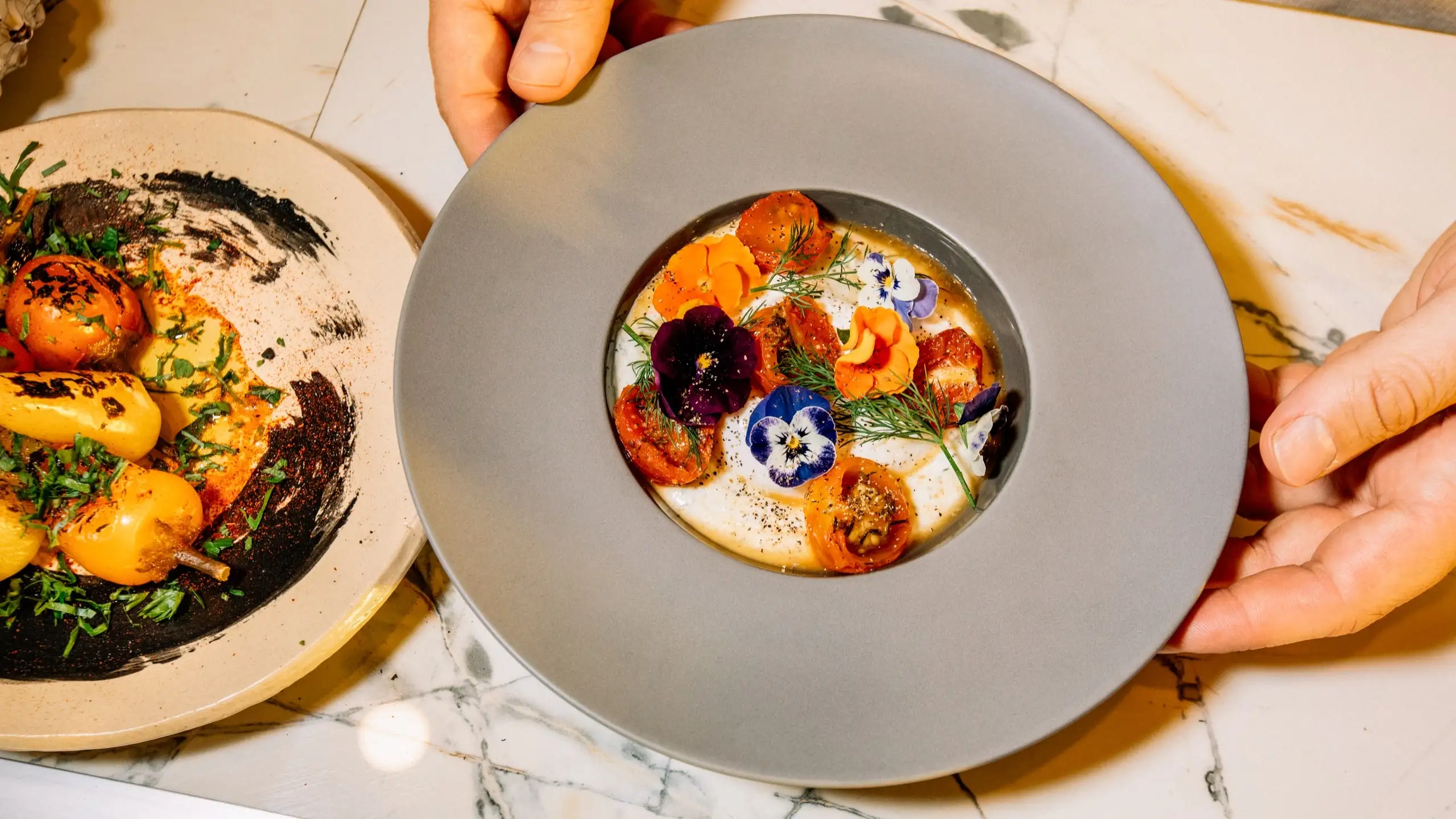 A dish of cheese dotted with flowers and herbs alongside a plate of peppers and eggplant.