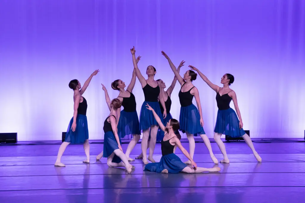 a group of female dancers wearing black leotards, blue ballet skirts, pink tights and shoes, posing on stage with a purple backdrop 