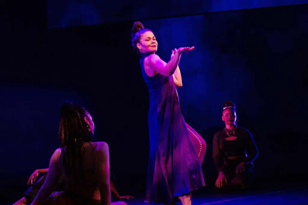 a female dancer on stage, other female dancers sitting around her, purple lighting 