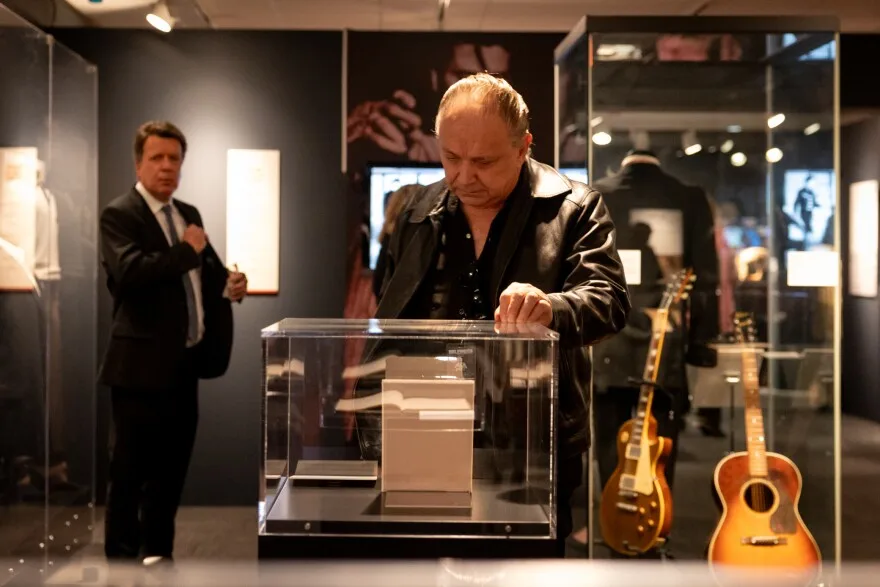 A man in a black dress shirt and black leather jacket looks down at an American music artifact encased in glass at the LBJ Presidential Library.