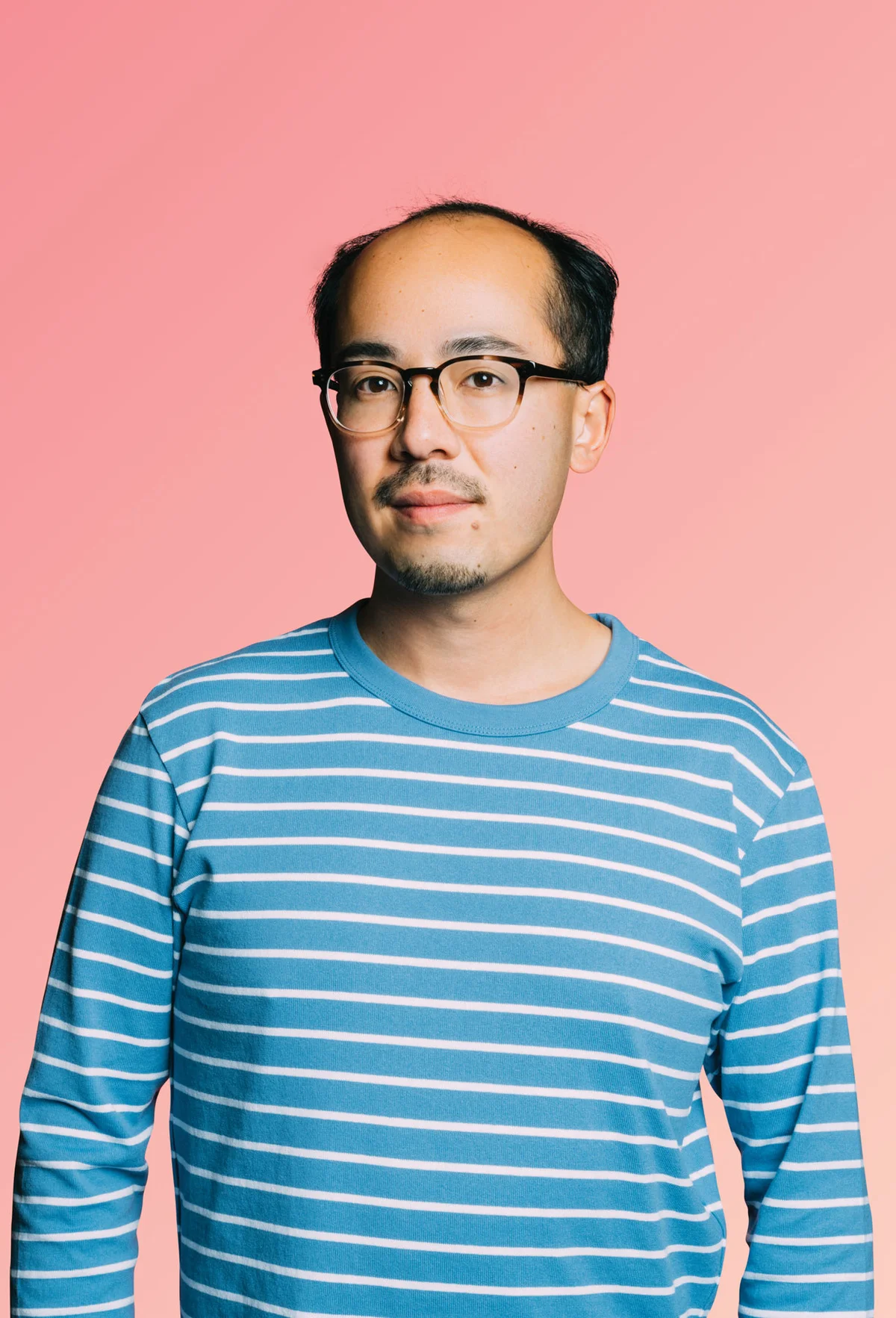 a headshot of a man with glasses and a goatee wearing a blue and white striped long sleeve shirt with a pink background