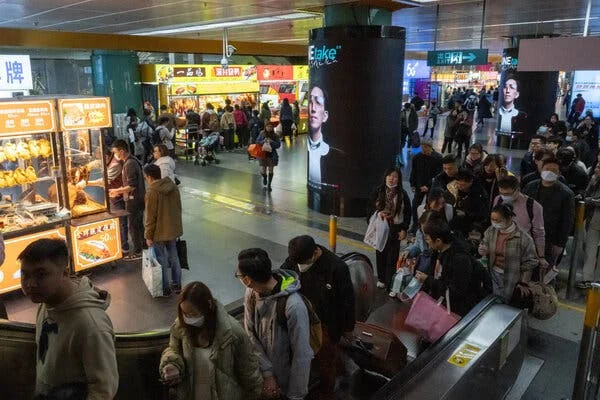 A crowd of people waiting to get on an escalator. Behind them are brightly lit shopping stalls. 