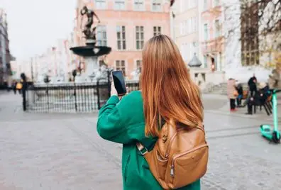 Happy cheerful young woman with phone walking on city street checks her smartphone. Traveling Europe in autumn. Gdansk, Poland