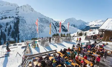 Skiers sit outside in the sunshine at long tables, eating and drinking. There are brightly coloured flags, and the restaurant looks out on to a view of mountains.