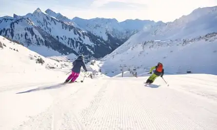 Two skiers in brightly-coloured gear ski downhill, towards the sun.