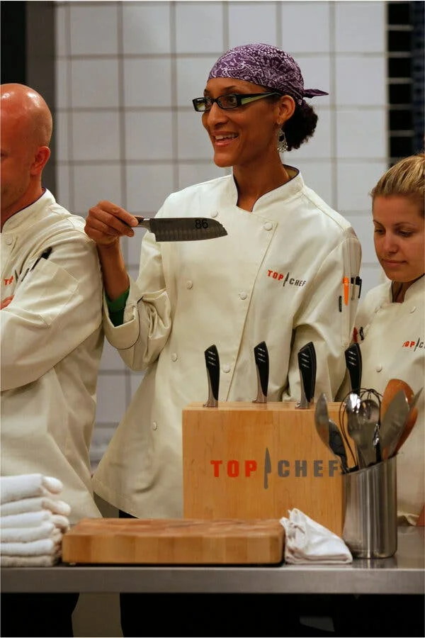 A tall woman in a chef’s coat and black glasses standing next to two other people in chef’s coats holds a knife.