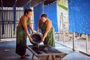 Ibu Sri and Ibu Muntiani prepare a bath for dyeing fabrics in one of SukkhaCitta’s craft schools, Rumah SukkhaCitta, East Java. The textiles hanging behind them have been decorated with a floral batik design.