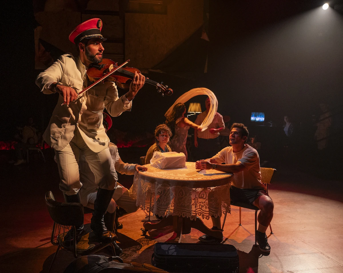 A soldier plays a violin standing on a chair next to two observers sitting at a table.