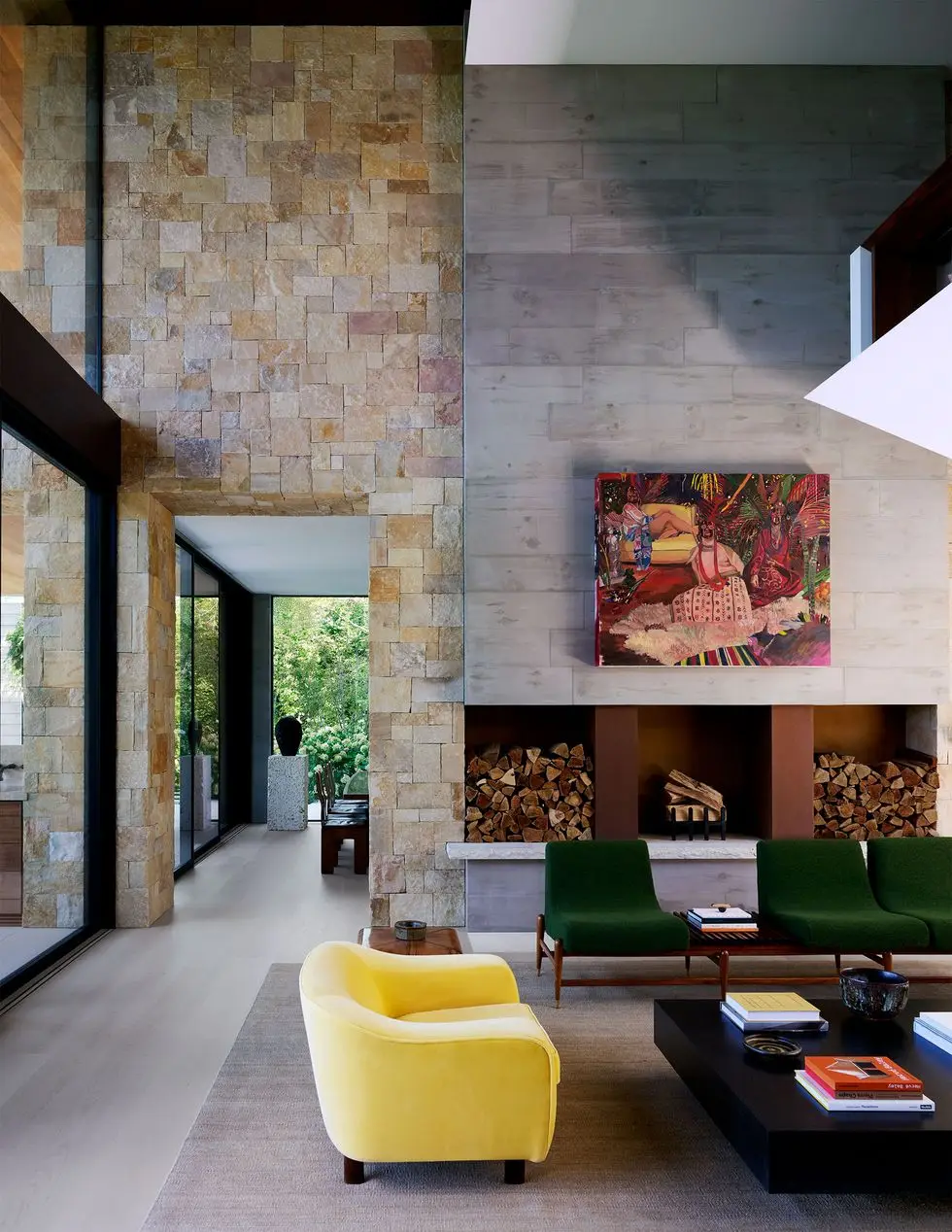 a living room with a partial cement wall and a partial stone wall, fireplace with niches filled with wood, a large painting above, green modular sofa with wood base, yellow loveseat, square cocktail table, taupe rug