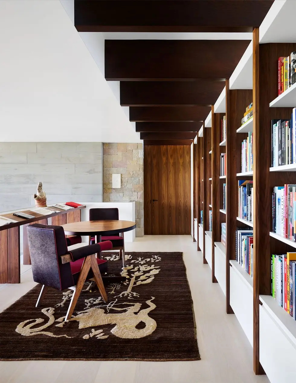 a mezzanine with a row of standing waist high shelves displaying open books, a small round table with two chairs, a wall of bookshelves with cabinets at bottom, brown rug with light colored abstract pattern