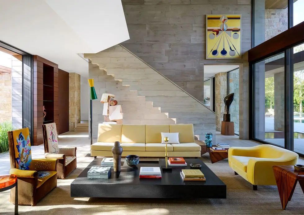 a living room with a cement wall and staircase, pale yellow and bright yellow sofas, two chairs upholstered in tapestries, large square cocktail table, side tables, floor lamp, sculptures and wall art, windows on two sides