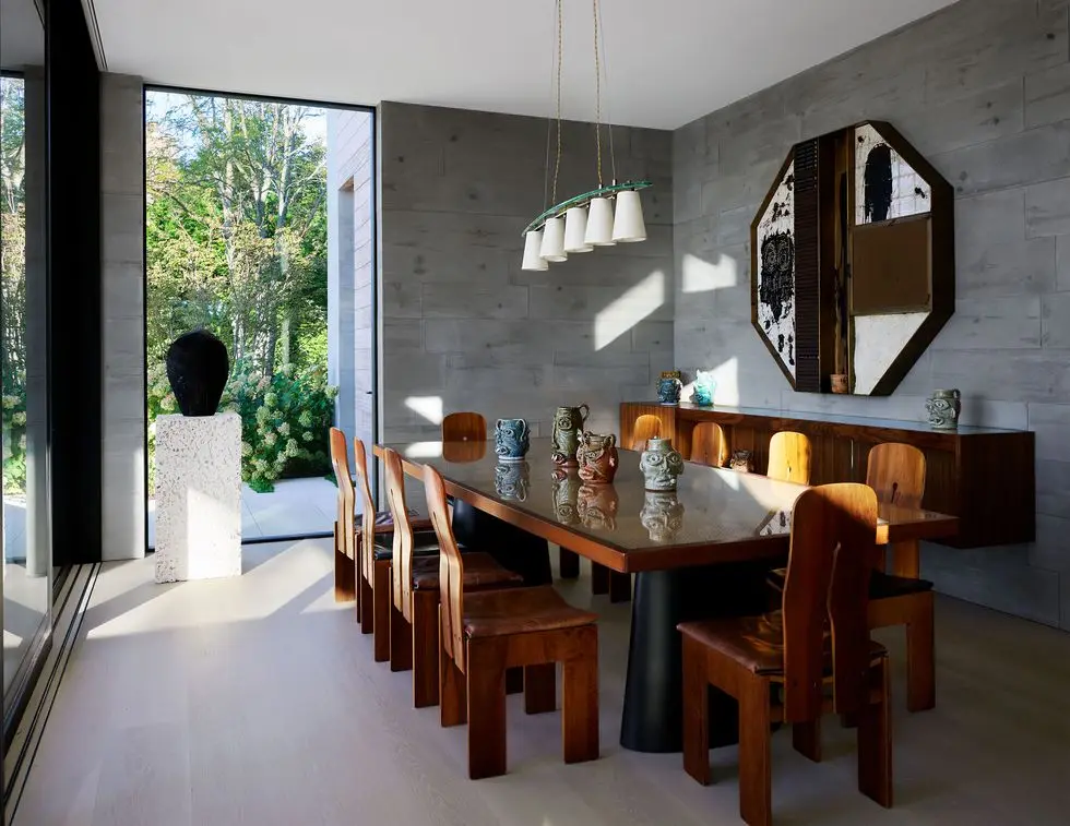 a dining room with long wooden table and 10 chairs with curved backs, ceramic pots with faces on table, five light pendant, floating credenza with hexagonal artwork above, sculpture on a plinth by windows