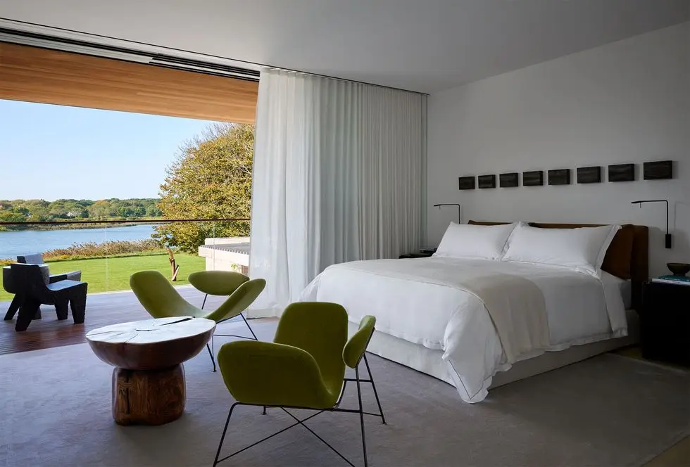 a bedroom with a glass wall opening onto a terrace overlooking a pond, white bedding on bed, dark headboard and nightstands, a row of small frames above bed, curved green armchairs, rustic wood table