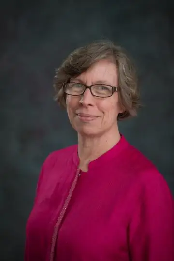 A headshot of Ann Miller, who has short hair and glasses and is wearing a magenta-colored shirt, looking at the camera against a grey backdrop.