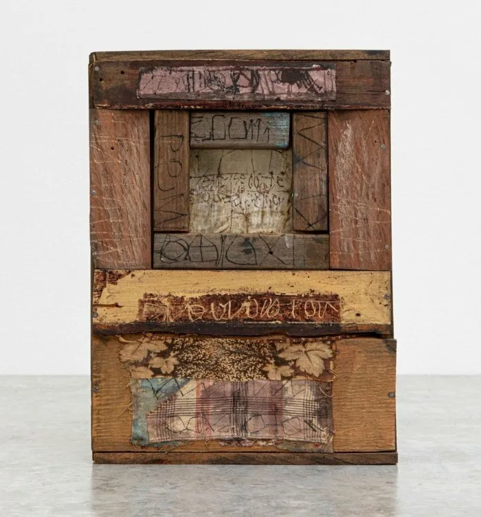 An artwork consists of a small wooden box with nails, fabric, paper printed and inked, and runic writing