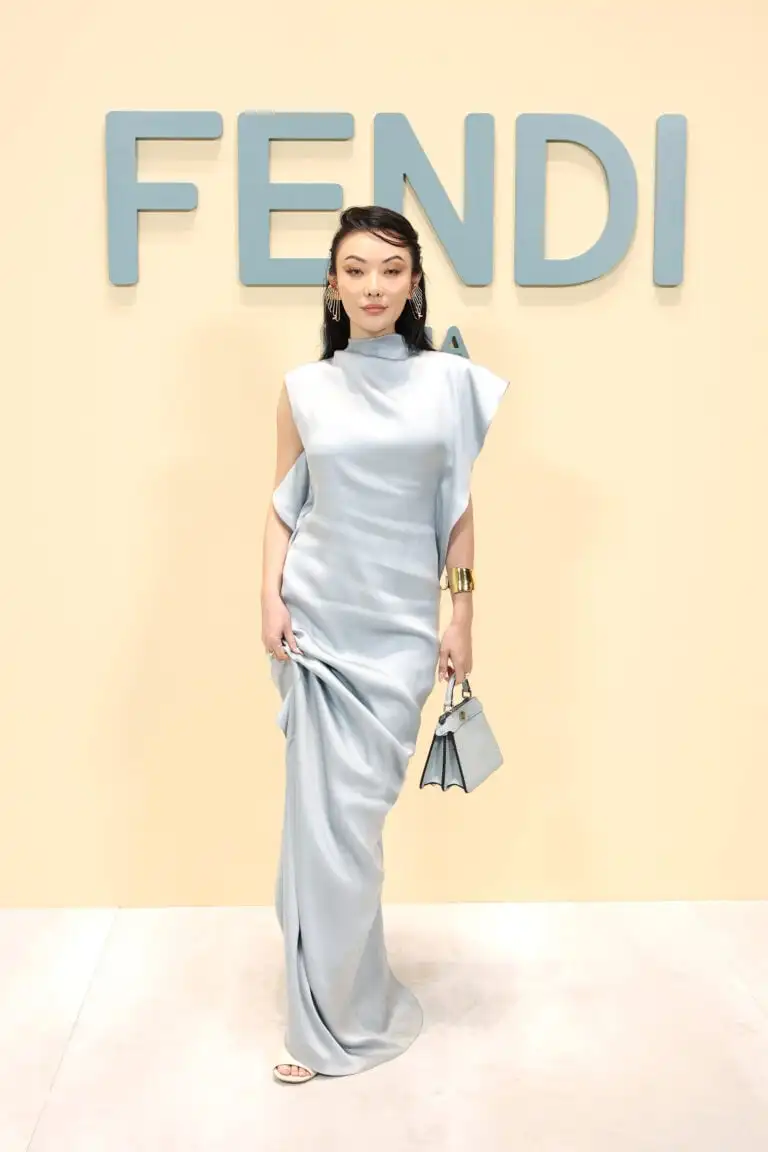 Jessica Wang stands poised against a Fendi backdrop at Milan Fashion Week. She models an elegant, light blue, high-collared, sleeveless gown that gracefully drapes to the floor, complete with sophisticated accessories including statement earrings, a bangle, and a petite two-tone handbag.